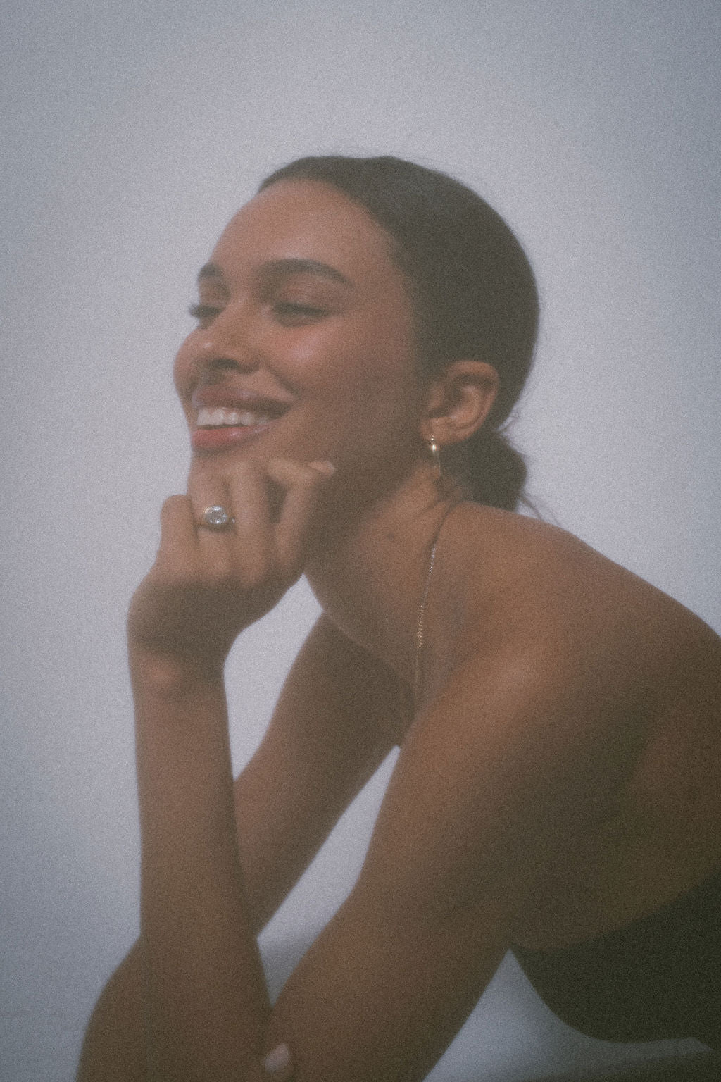 A smiling woman with dark hair tied back poses in a minimalist setting, wearing a delicate gold chain necklace and a gold ring with a clear gemstone.