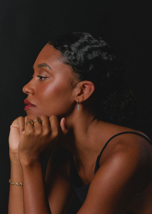 A woman in profile wearing delicate gold rings and a gold bracelet against a dark black background. (view 2)