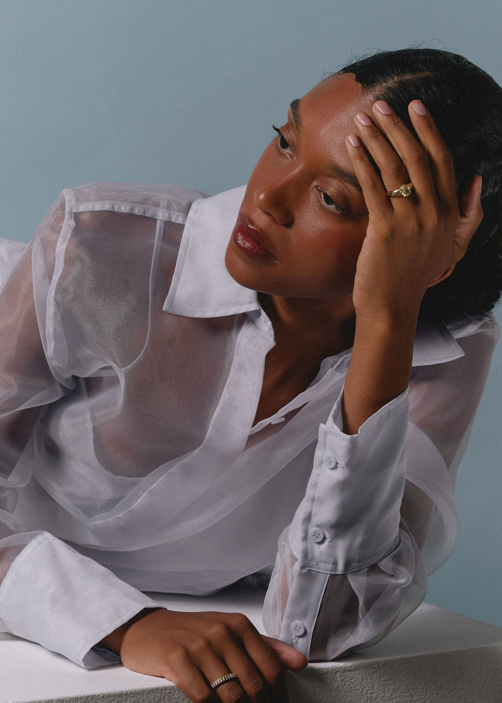 A woman wearing a sheer white button-up shirt, resting her head on her hand against a solid light blue background. (view 11)