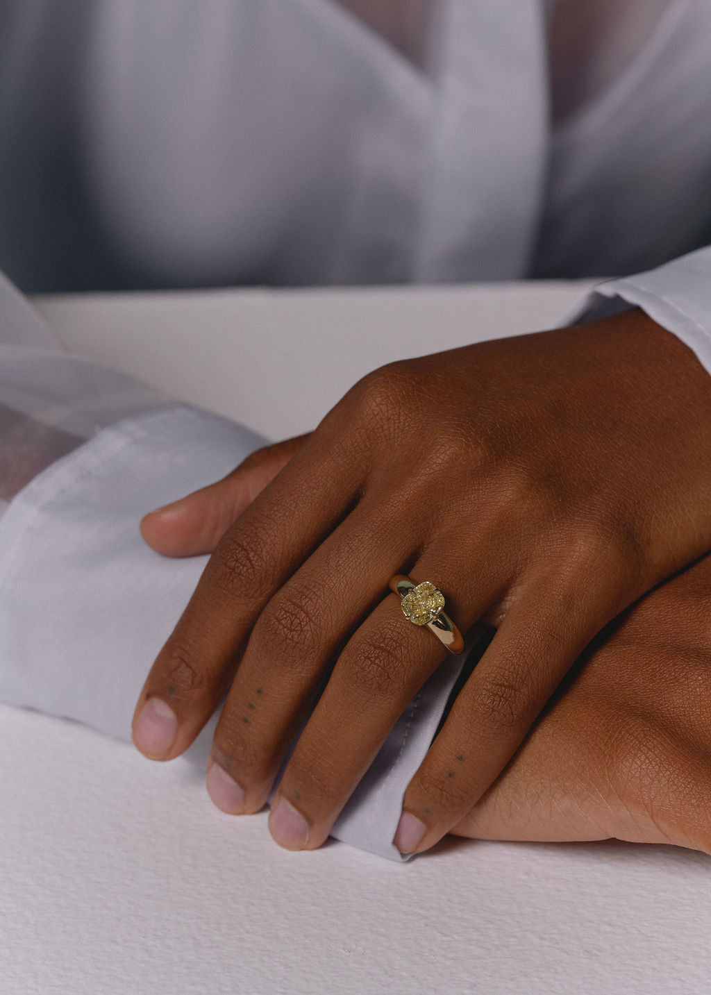 A close-up of a person's hand wearing a yellow gold ring with a yellow diamond cushion center resting on a white surface. (view 10)
