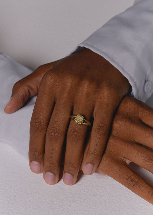 A close-up of a person's hand wearing a small gold ring with a yellow diamond center stone, resting on a white textured surface. (view 2)