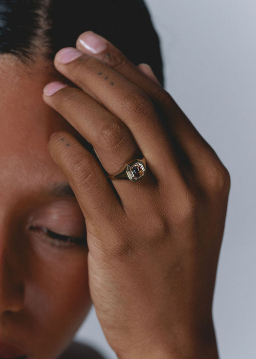 A close-up lifestyle shot of a person wearing a gold signet ring with a clear rectangular gemstone on their finger, with their hand resting against their face. (view 3)