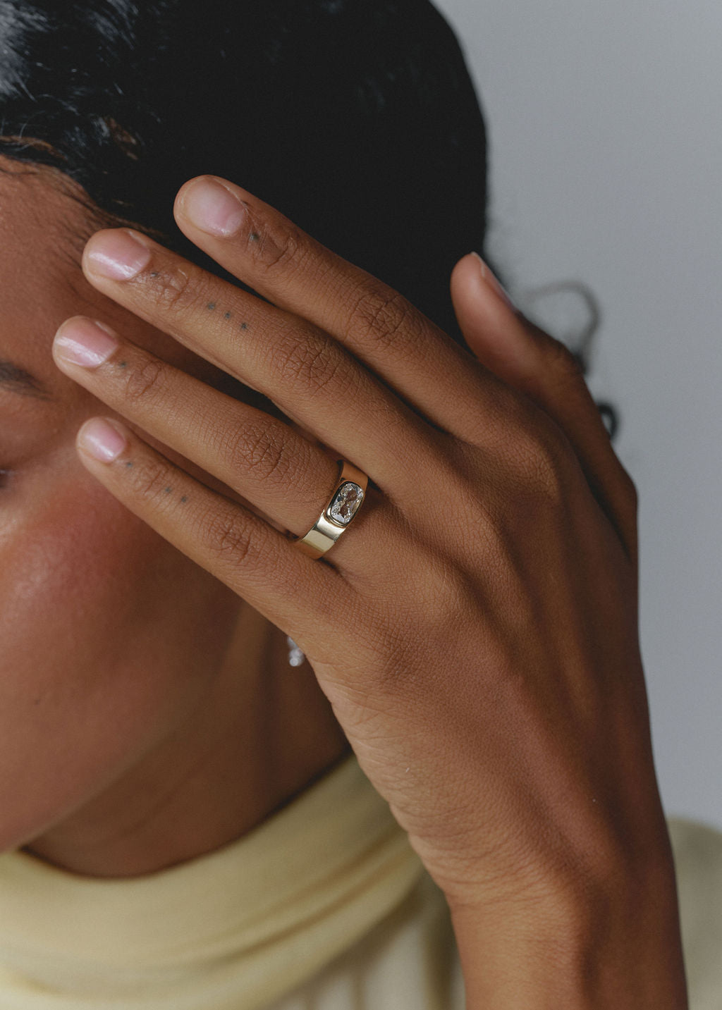 A close-up lifestyle shot of a person wearing a gold band ring with a clear rectangular gemstone on their ring finger, with their hand resting against their face. (view 2)