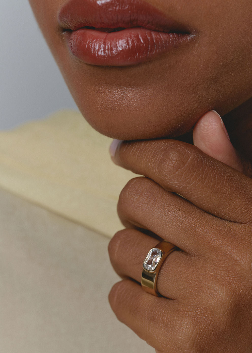 A close-up lifestyle shot of a person's hand resting on their chin, wearing a gold ring with a rectangular clear gemstone on the ring finger. (view 3)
