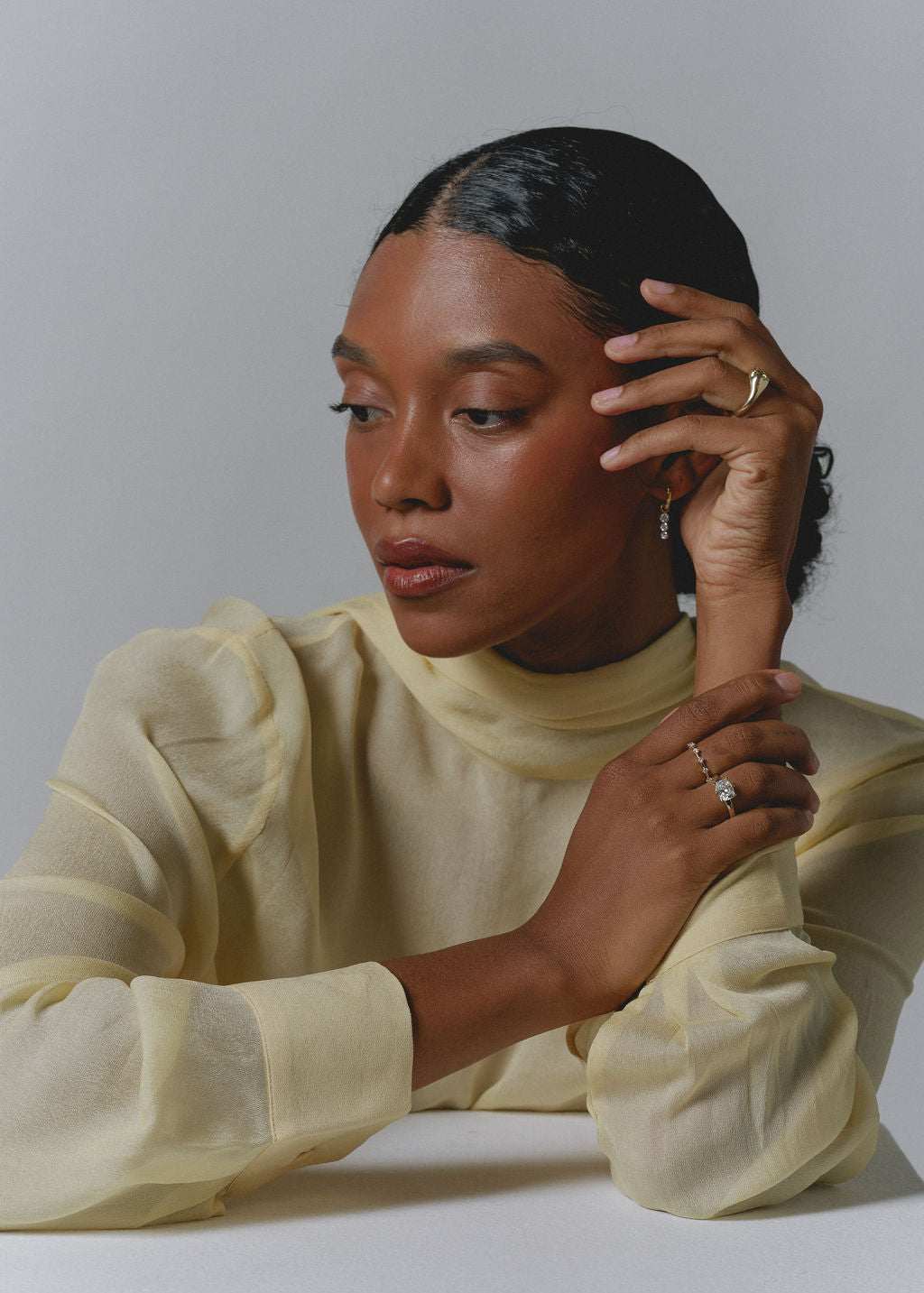 A woman with dark hair pulled back wears a pale yellow sheer long-sleeve mock-neck top and gold rings, posing against a light gray studio background. (view 7)