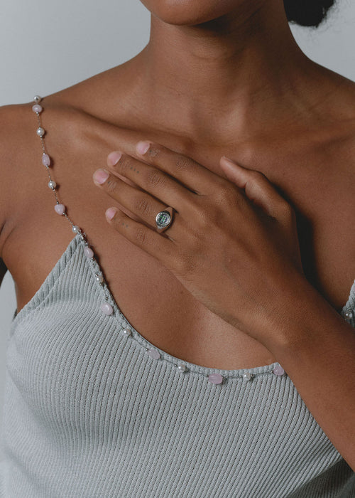 A close-up lifestyle shot of a model wearing a silver-toned ring with a round dark stone, paired with a ribbed light gray top and a delicate beaded necklace. (view 3)