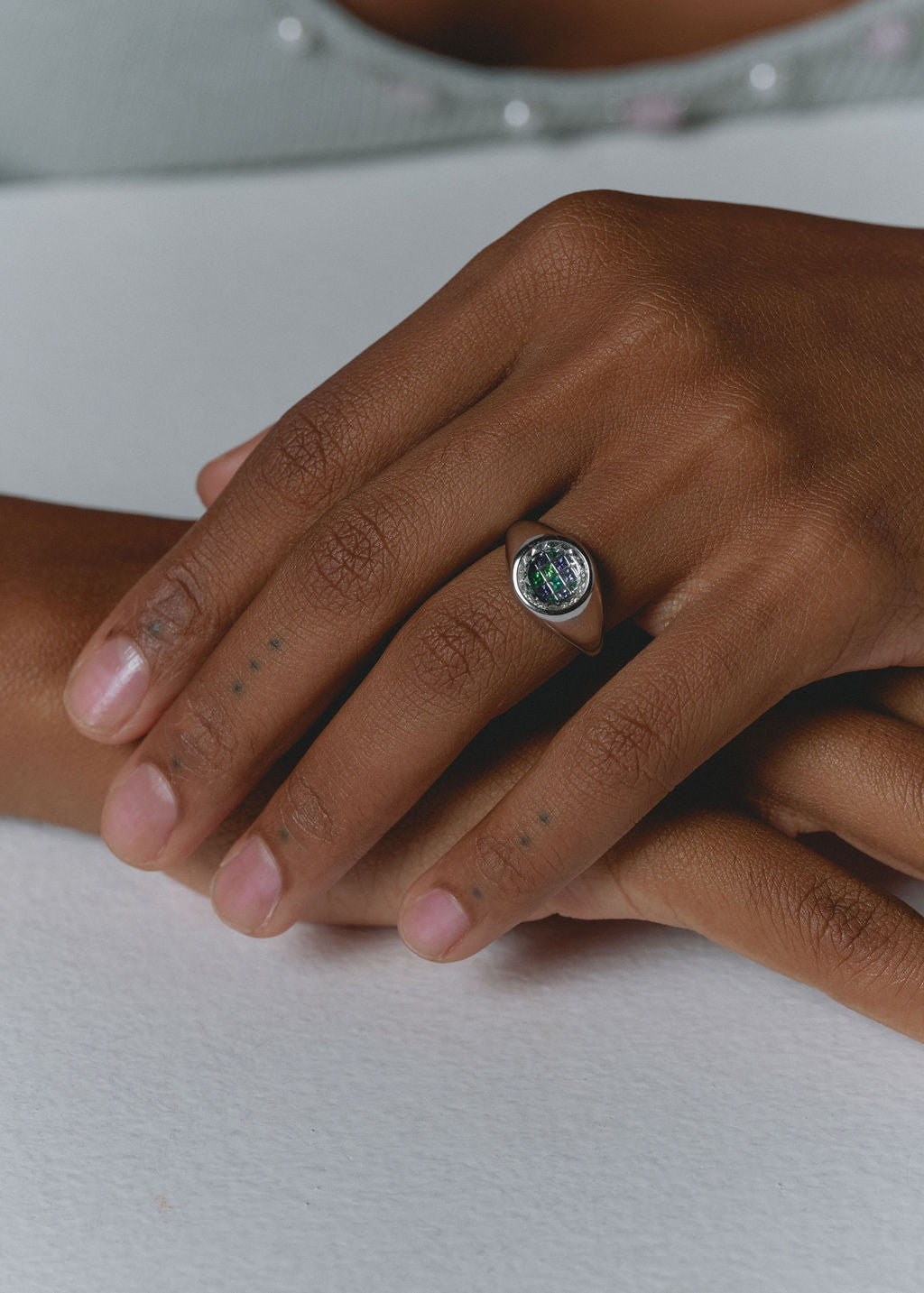 A close-up of a person's hands resting on a white surface, wearing a silver signet ring with a blue and green gemstones. (view 2)
