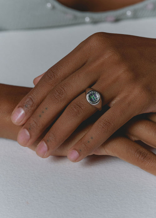 A close-up of a person's hands resting on a white surface, wearing a silver signet ring with a blue and green gemstones. (view 2)