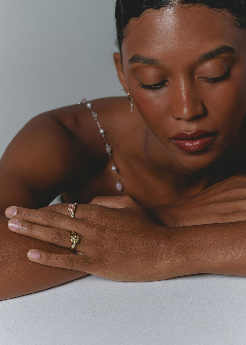 A close-up lifestyle shot of a woman, resting her arms on a light gray surface. She is wearing gold rings on her fingers and has a delicate pearl strap over her shoulder. (view 4)