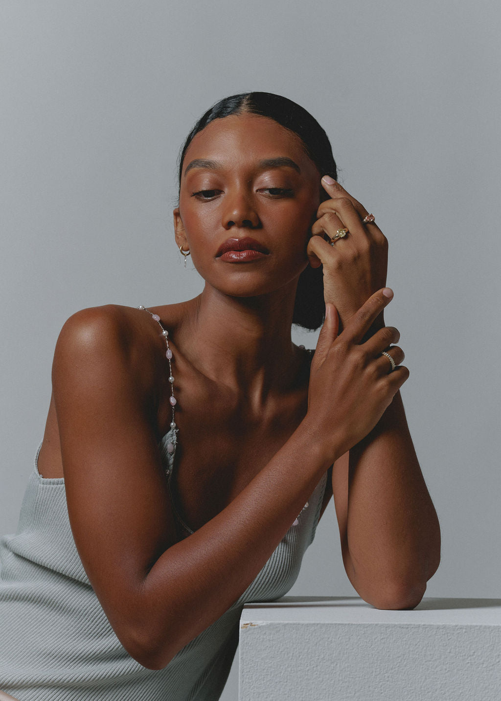 A woman with dark hair pulled back wears a light gray ribbed camisole and delicate gold rings, posing against a neutral gray background. (view 8)