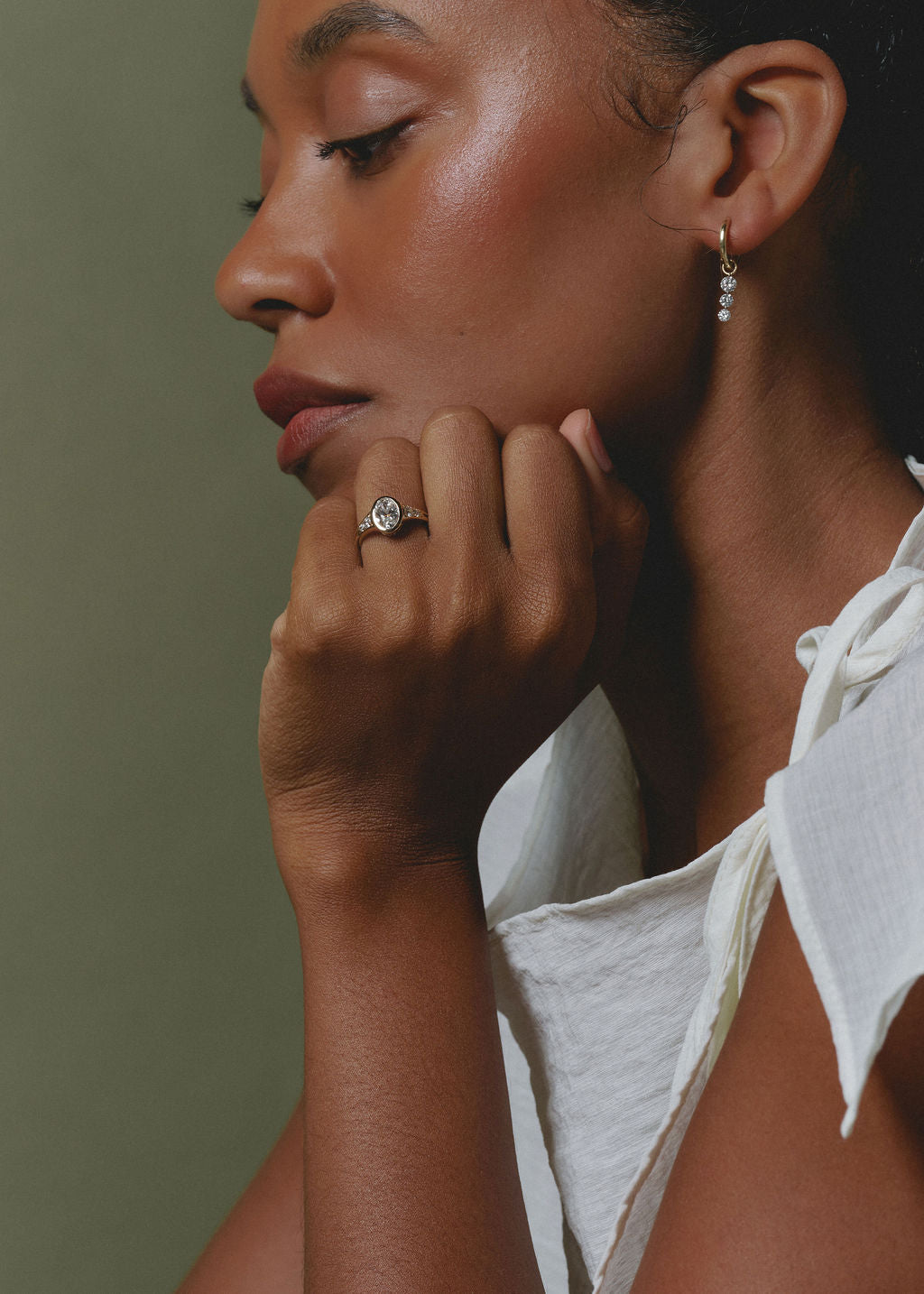 A close-up portrait of a woman wearing a silver ring with a clear round gemstone and a small silver hoop earring with clear stones, posed against a muted olive green background.