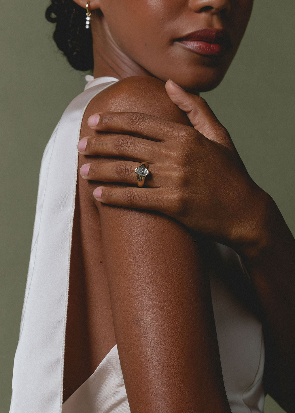 A close-up lifestyle shot of a model wearing a gold ring with a pear-cut clear gemstone on her finger, resting her hand on her shoulder. She is wearing a white sleeveless top against a muted olive green background. (view 4)