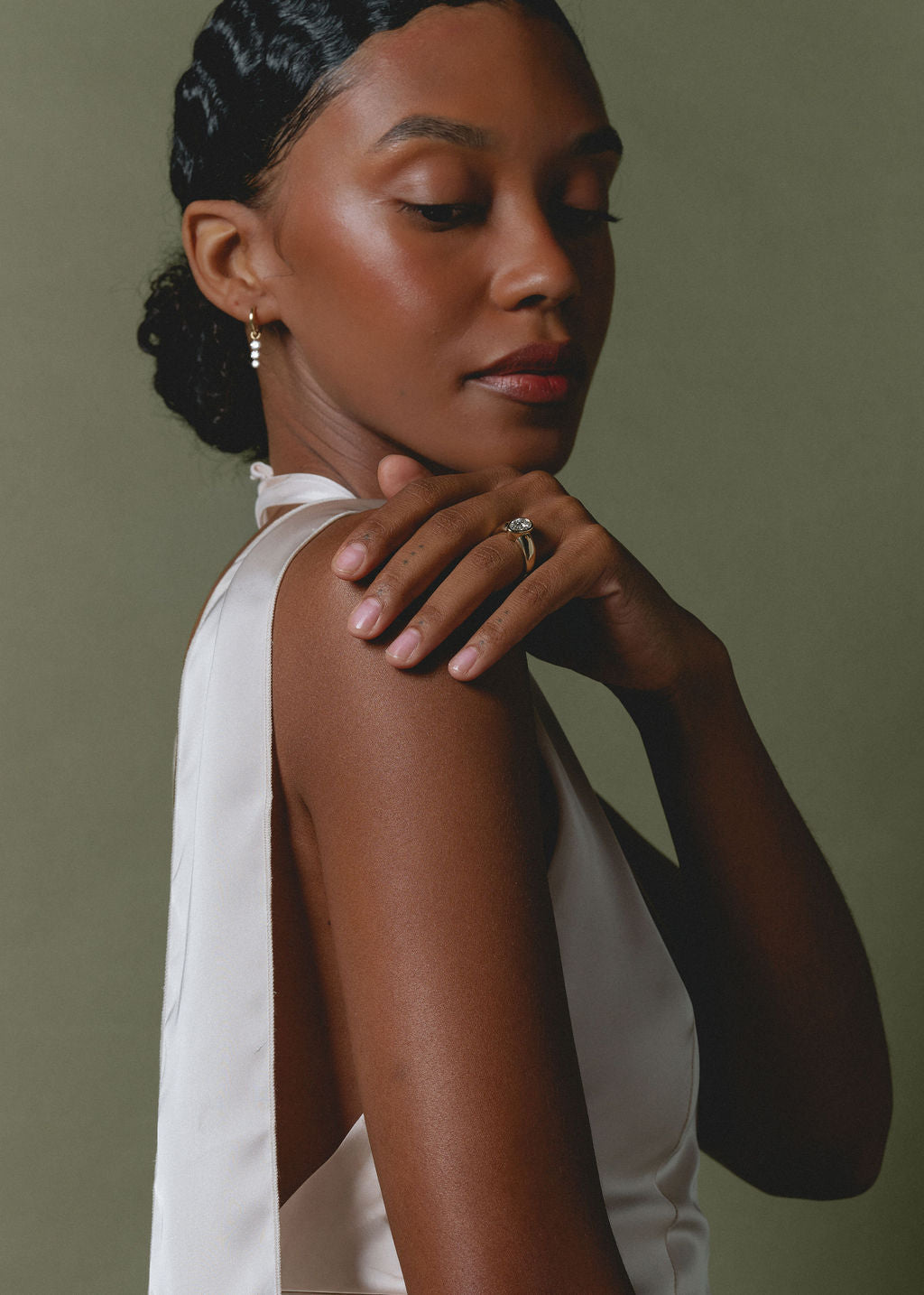 A woman with hair pulled back wears a white sleeveless top and delicate gold jewelry, including a ring and earring, posing against a muted olive green background. (view 2)