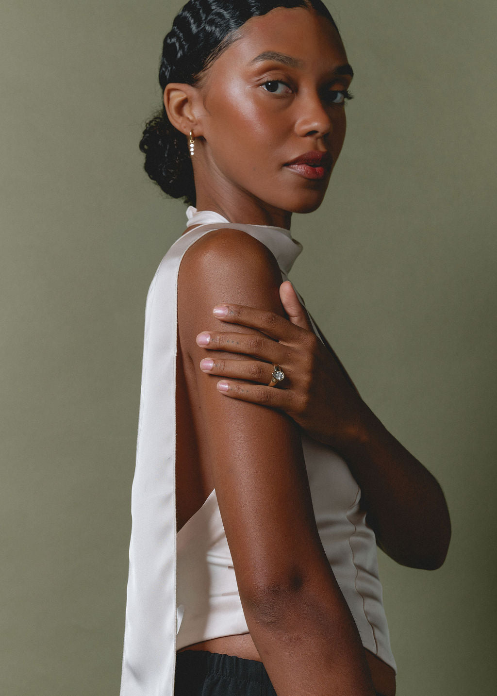 A woman with her hair pulled back wears a white sleeveless top with a draped side detail and a high neckline, posed against a muted olive-green background. She is wearing a ring on one finger. (view 5)