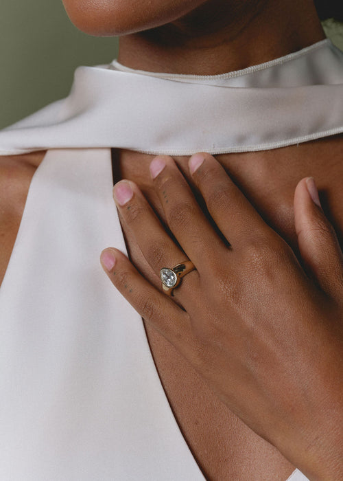 A close-up lifestyle shot of a woman wearing a gold ring with a pear cut clear gemstone on her finger, resting her hand on her chest. She is wearing a white satin high-neck top.
