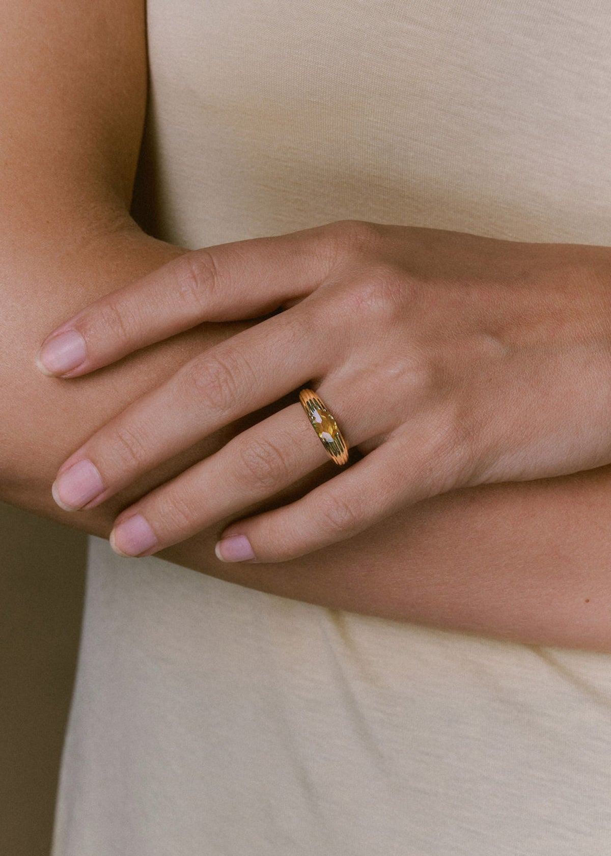 A close-up of a person's hand wearing a ridged gold band ring with a yellow sapphire center, set against a soft beige sleeveless top. (view 3)