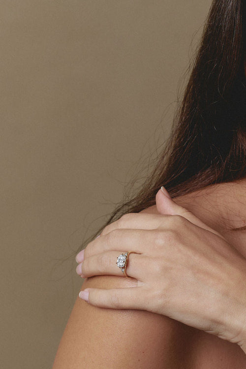 A close-up lifestyle shot of a woman wearing a silver-toned diamond engagement ring on her finger, resting her hand on her shoulder against a solid beige background. (view 4)