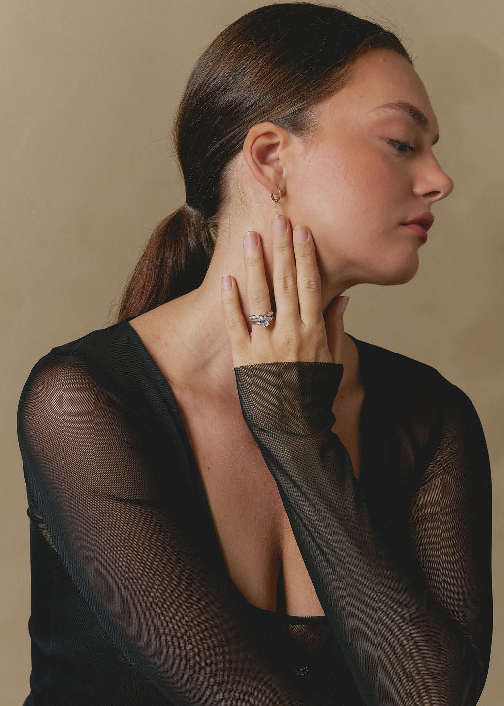 A woman in profile wearing a sheer black long-sleeve top, showcasing a delicate gold hoop earring and a silver-toned ring with a clear gemstone on her finger, set against a warm beige background. (view 11)