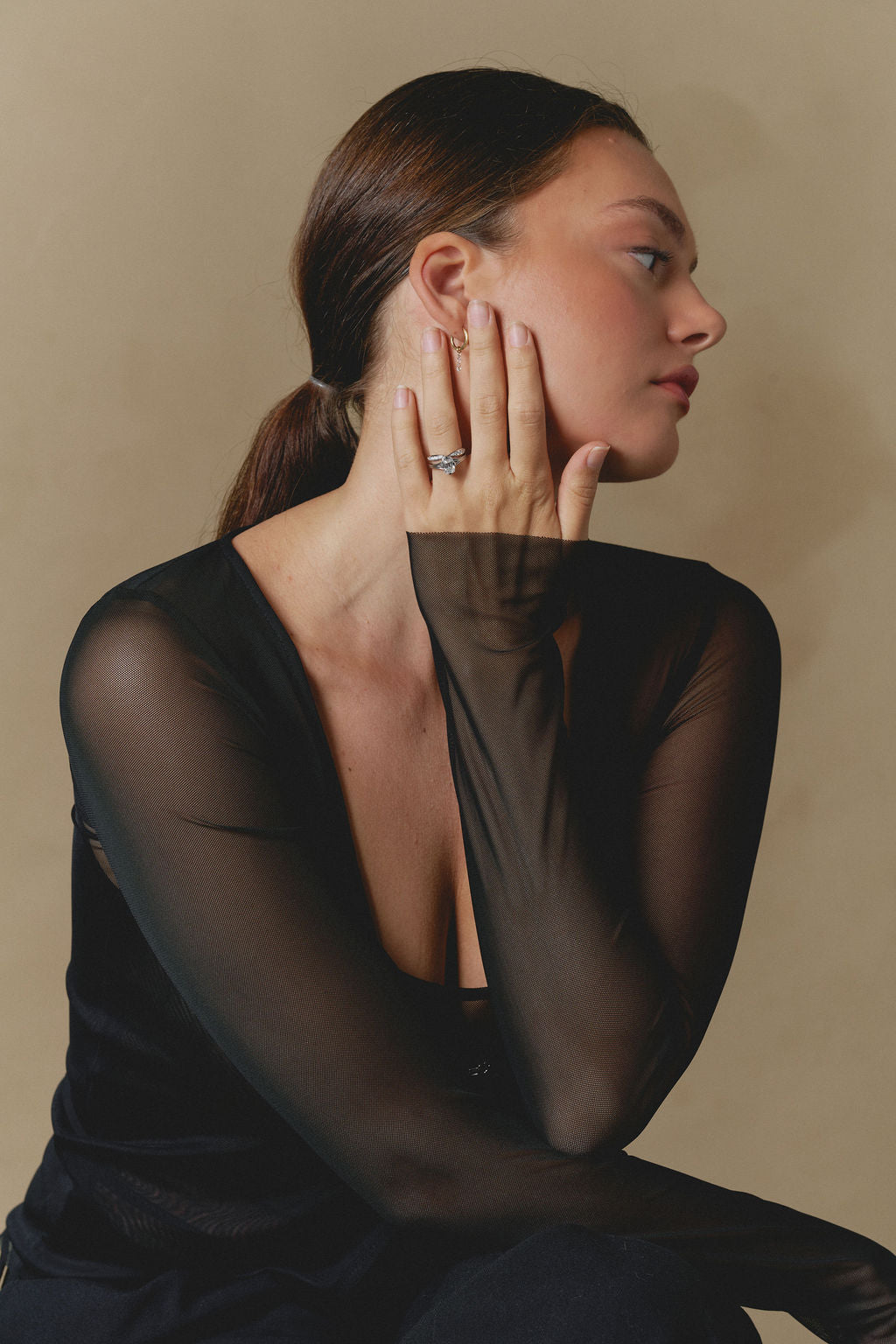 A woman in a black sheer long-sleeve top poses in profile against a warm beige background, wearing a delicate white gold ring on her finger. (view 7)