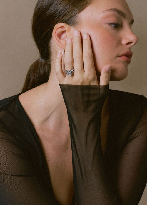 A close-up portrait of a woman wearing a silver ring with a clear central gemstone on her finger, paired with a sheer black top against a neutral beige background. (view 5)