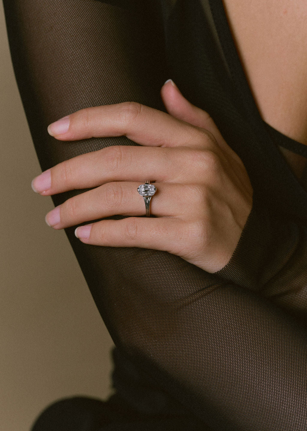 A close-up of a hand wearing a white gold solitaire ring with a elongated cushion diamond, set against a dark sheer black top and a warm beige background. (view 3)