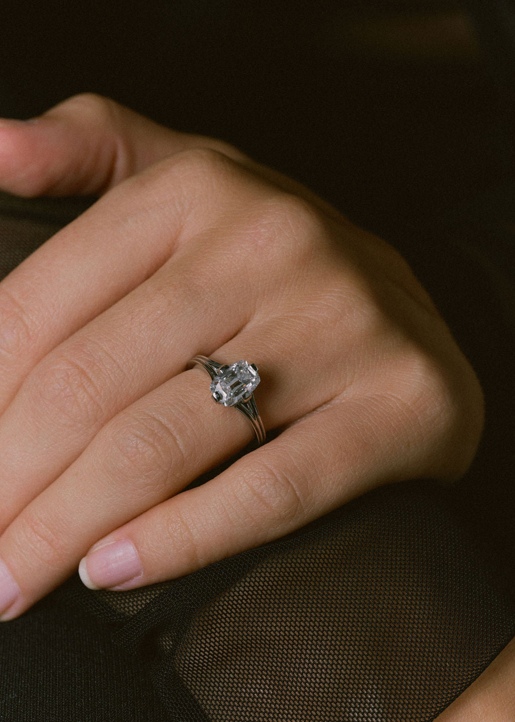 A close-up of a hand wearing a silver-toned ring with a large clear gemstone, set against a dark brown textured background.