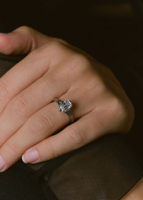 A close-up of a hand wearing a silver-toned ring with a large clear gemstone, set against a dark brown textured background.