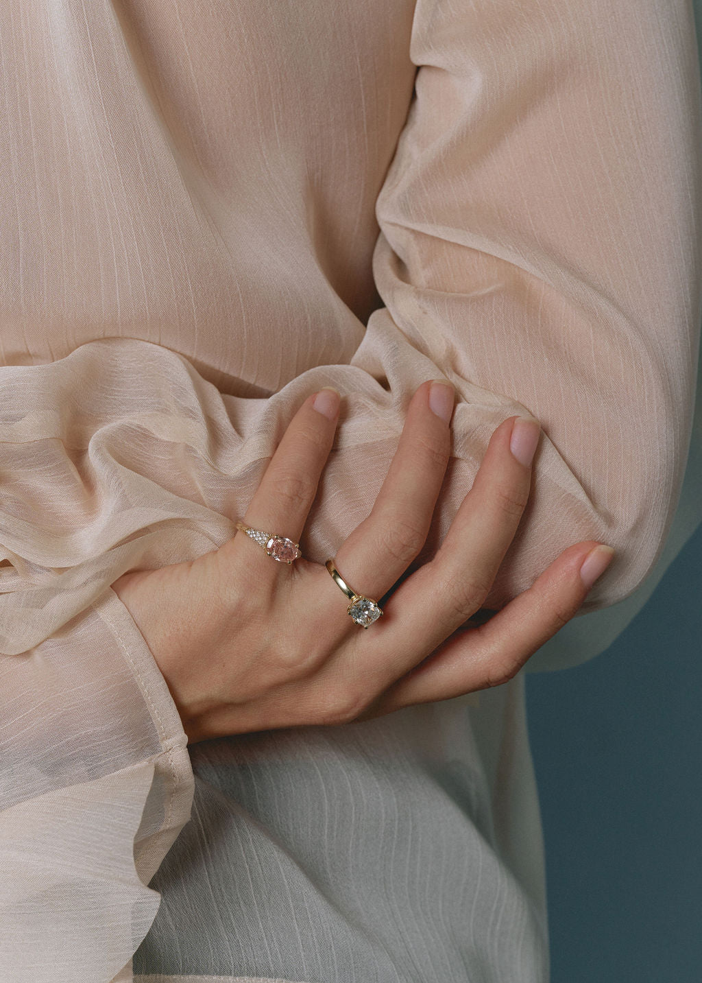 A close-up of a person's hand wearing two delicate rings, one with a pink stone and one with a clear center stone on a gold band, styled with a sheer, ruffled champagne-colored blouse against a muted blue-gray background. (view 5)