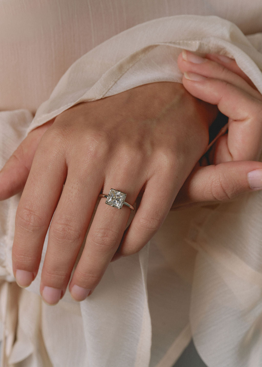 A close-up of a hand wearing a silver-toned ring with a square-cut clear gemstone, styled with soft cream-colored fabric. (view 11)
