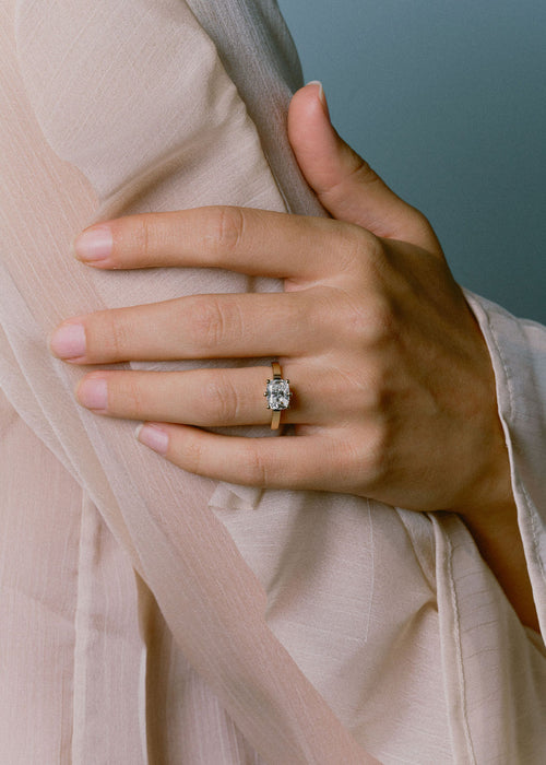 A close-up of a hand wearing a gold ring with a cushion cut diamond, resting on soft beige fabric against a muted blue-gray background. (view 2)