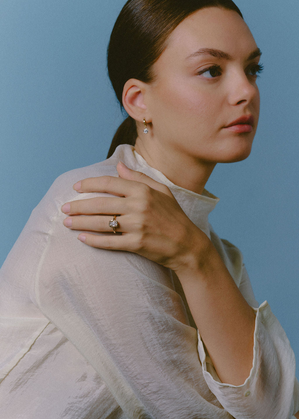A woman in a white high-neck blouse poses against a solid blue background, wearing a delicate gold ring with a champagne central stone on her finger and a matching small gold hoop earring with a dangling diamond. (view 11)