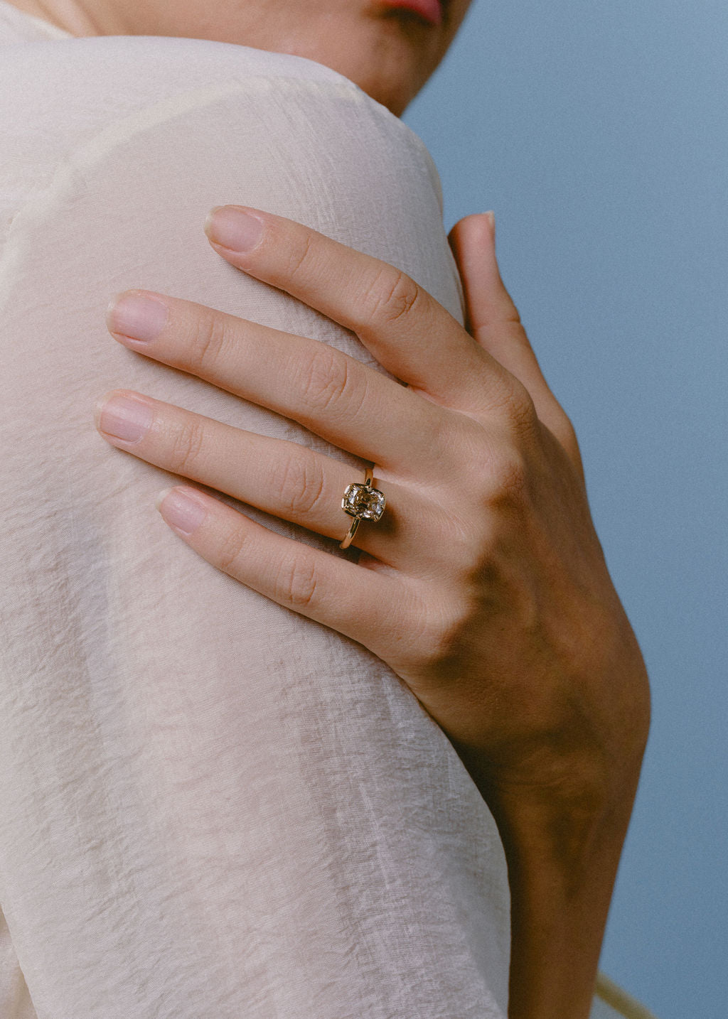 A close-up of a person's hand resting on their shoulder, wearing a gold ring with a champagne cushion gemstone. The person is wearing a light-colored, semi-sheer top against a solid light blue background. (view 8)