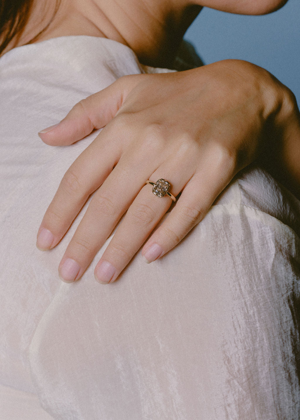 A close-up of a person's hand resting on their chest, wearing a delicate gold ring with a small, champagne cushion gemstone. (view 6)