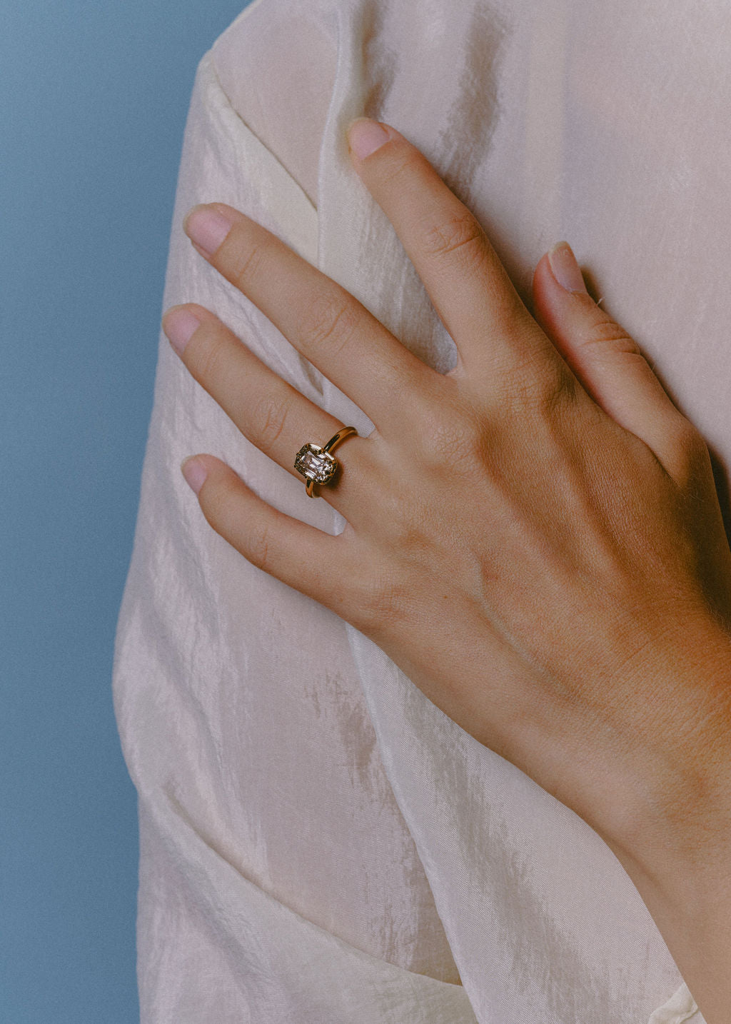 A close-up of a person's hand resting on a textured cream fabric, wearing a gold ring with a champagne gemstone against a solid blue background. (view 12)