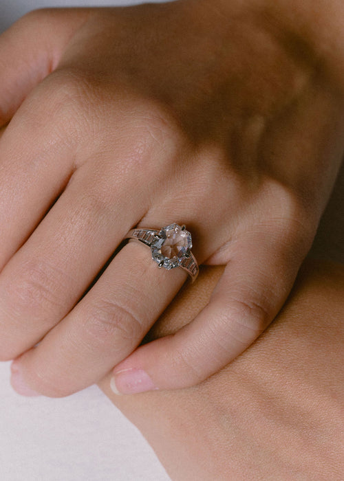 A close-up of a hand wearing a silver-toned ring with a white diamond center stone, resting on another hand against a soft light background.