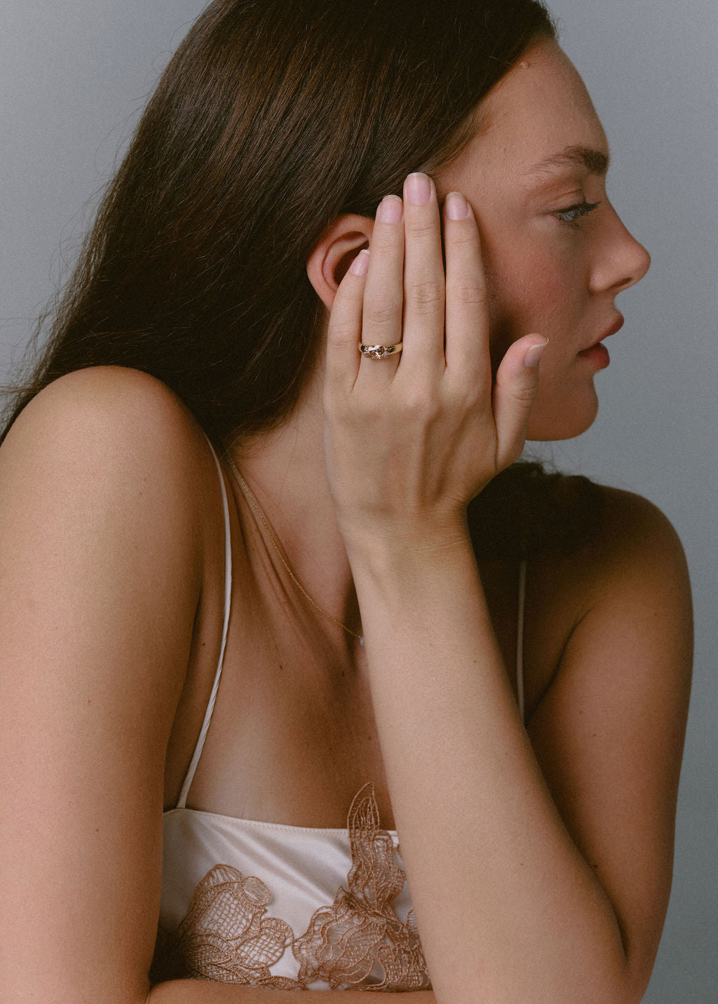 A woman in profile wearing a gold ring on her finger, posing in a white lace-trimmed camisole against a solid gray background. (view 11)