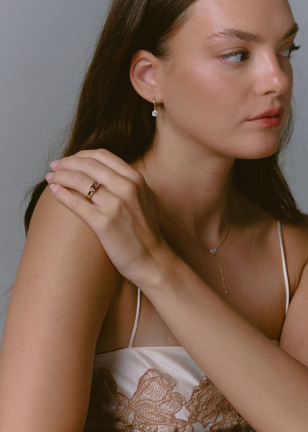 A close-up lifestyle shot of a woman wearing delicate gold jewelry, featuring a gold ring on her finger set against a plain gray background. (view 5)