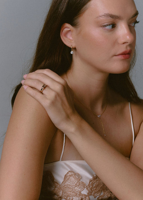 A close-up lifestyle shot of a woman wearing delicate gold jewelry, featuring a gold ring on her finger set against a plain gray background. (view 5)