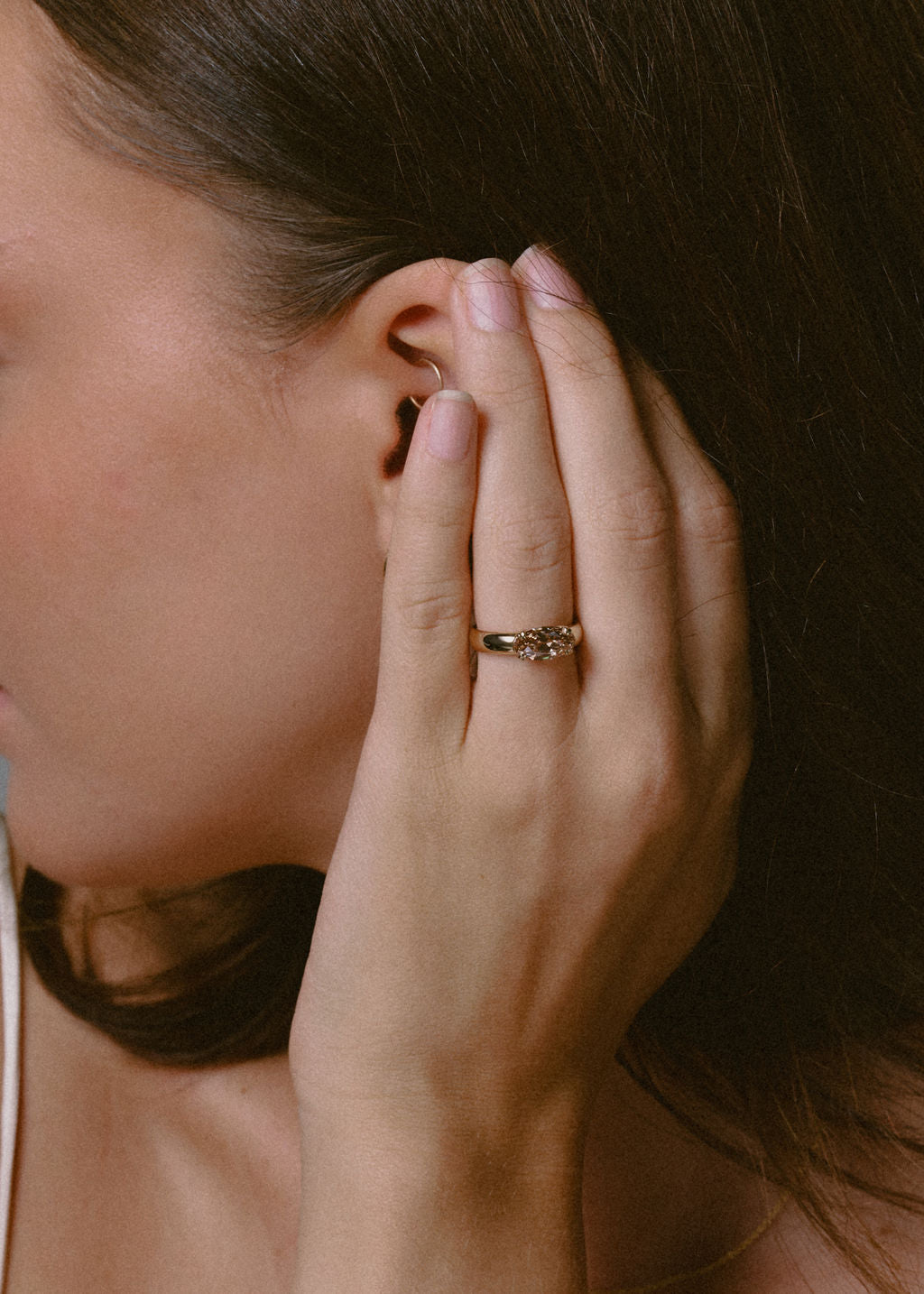 A close-up lifestyle shot of a woman's ear and hand wearing a gold ring with a champagne gemstone on her ring finger. (view 4)