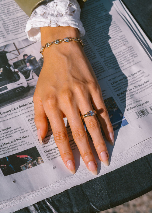 A close-up lifestyle shot of a woman's hand resting on a newspaper, showcasing a gold chain bracelet on the wrist and a gold ring with ca single diamond on her finger. (view 3)