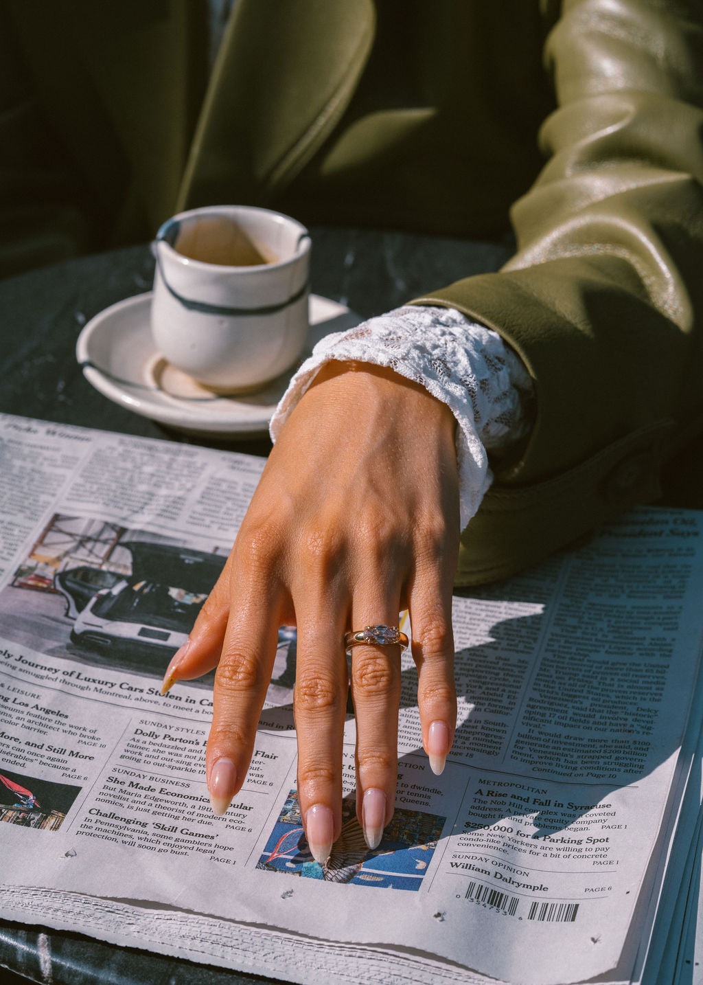 A ifestyle shot of a person's hand resting on an open newspaper, wearing a gold ring with a central gemstone. The person is wearing an olive green leather jacket and a white lace cuff. (view 10)