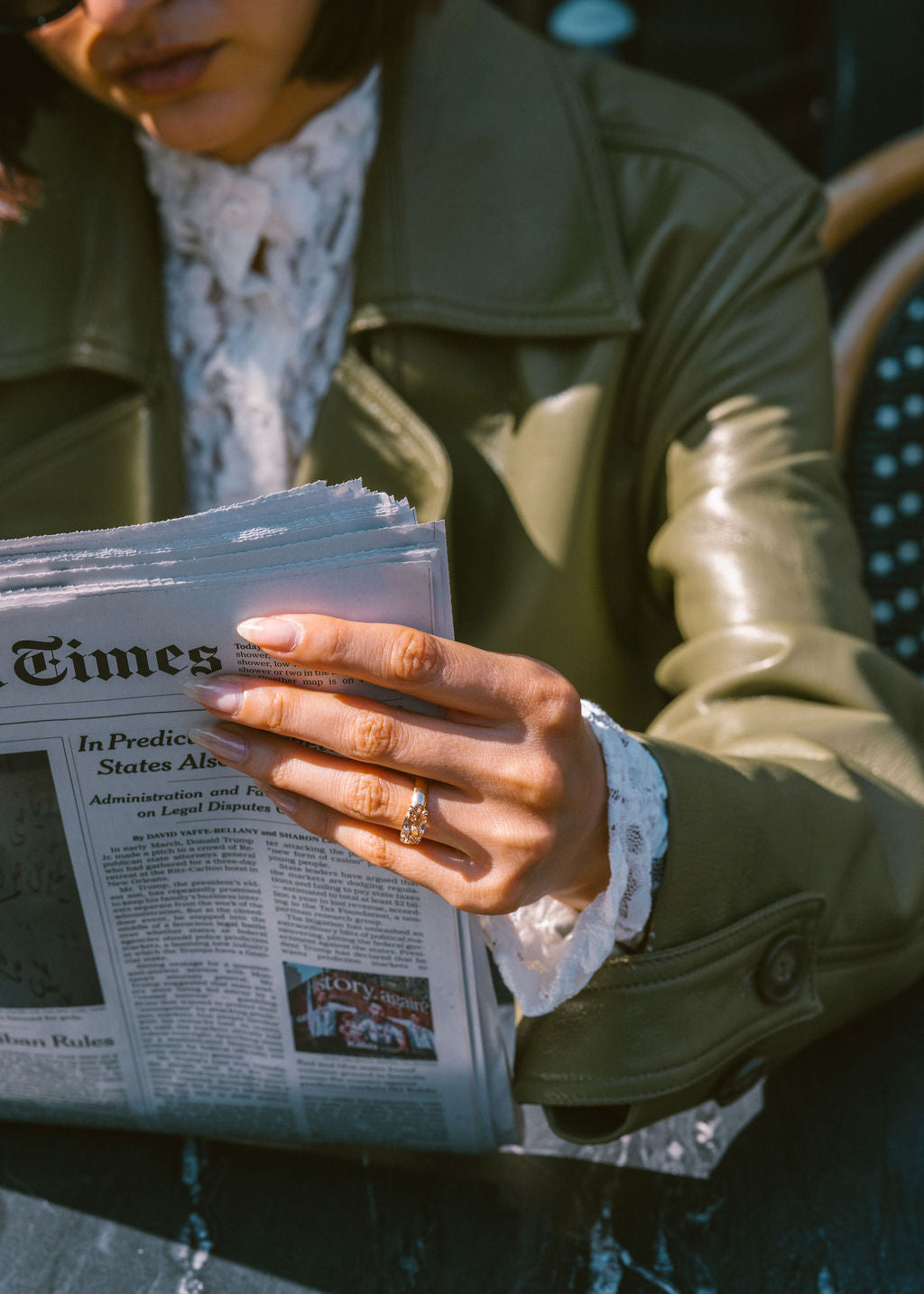A close-up lifestyle shot of a person wearing an olive green leather jacket and a white lace shirt, holding a newspaper while wearing a ring on their finger. (view 7)
