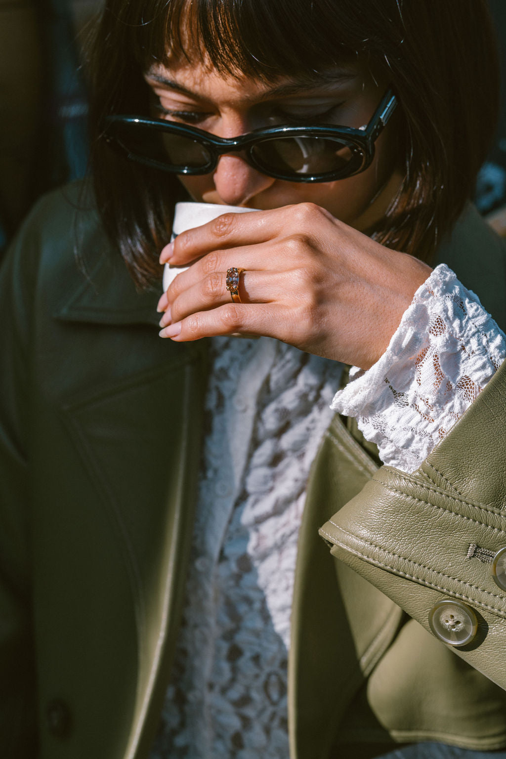 A close-up lifestyle shot of a person wearing black cat-eye sunglasses, a green leather jacket, and a white lace top, holding a white paper coffee cup. A gold ring is visible on their finger. (view 8)