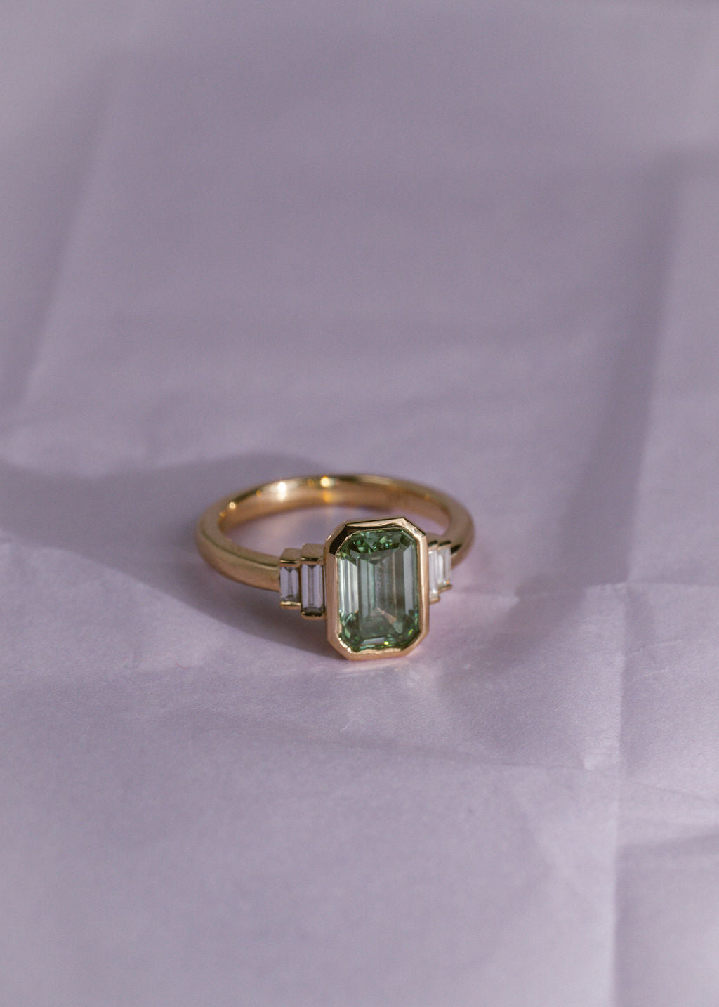 A yellow gold ring with a green diamond emerald center stone and white diamond side stones on a pale purple background.