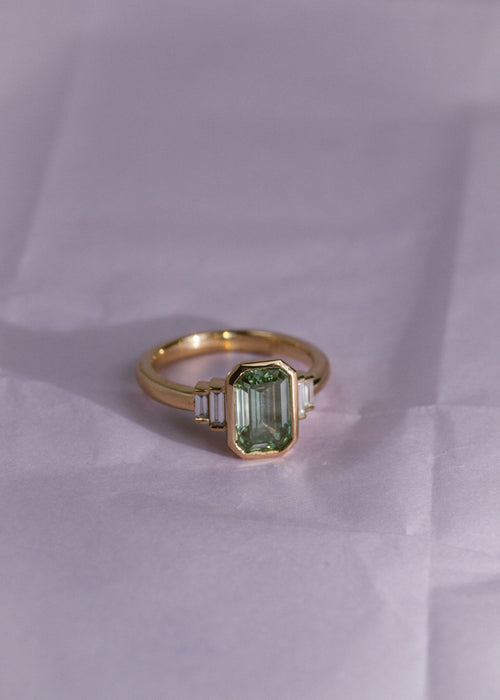 A yellow gold ring with a green diamond emerald center stone and white diamond side stones on a pale purple background.