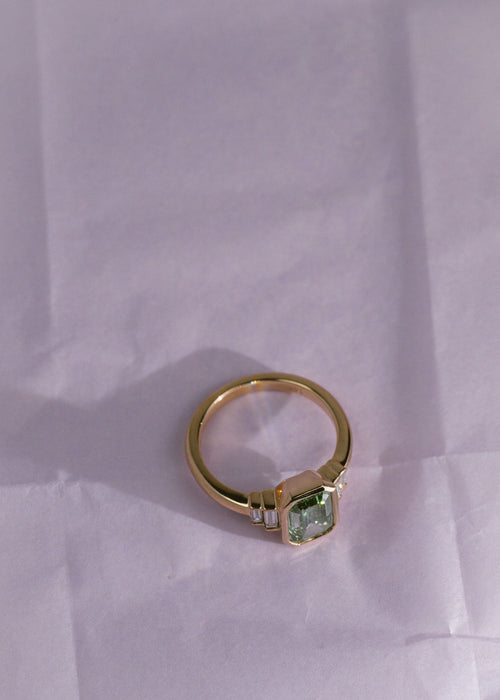 A yellow gold ring with a green diamond emerald center stone and white diamond side stones on a pale purple background. (view 4)