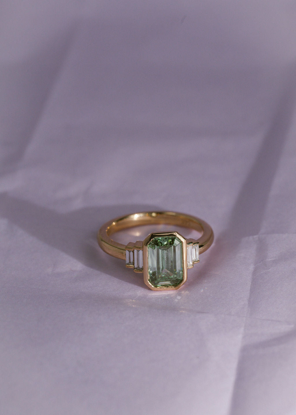 A yellow gold ring with a green diamond emerald center stone and white diamond side stones on a pale purple background. (view 7)