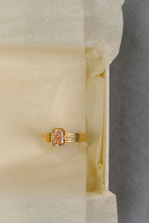 A gold ring with a rectangular pink diamond, displayed on a piece of cream-colored tissue paper. (view 2)