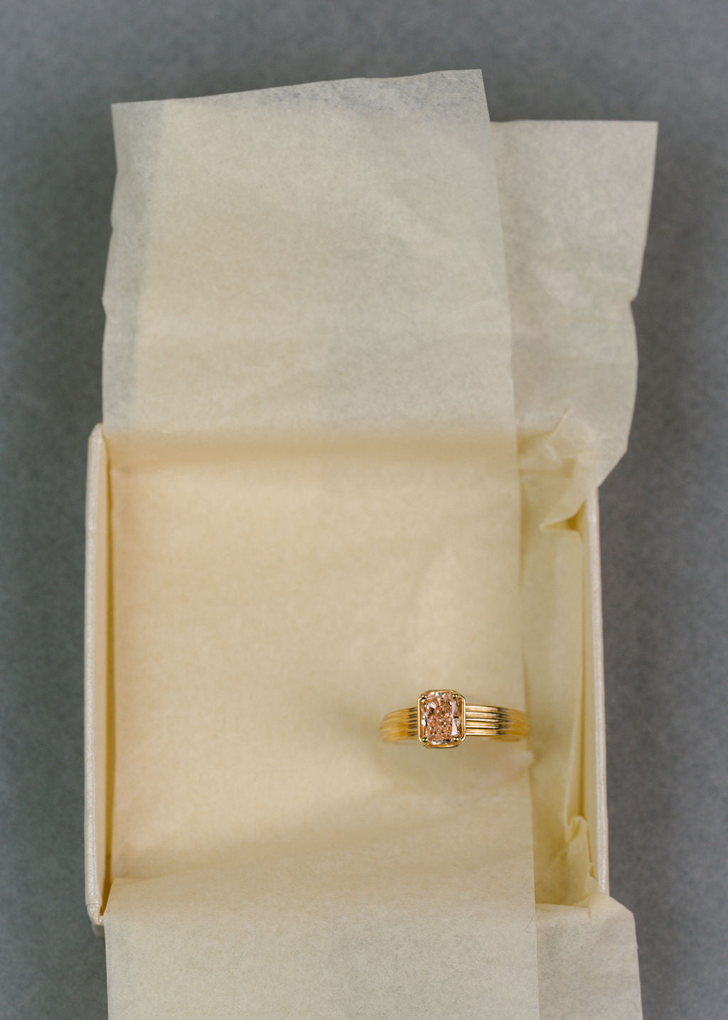 A gold ring with a rectangular pink diamond, displayed on a piece of cream-colored tissue paper. (view 5)