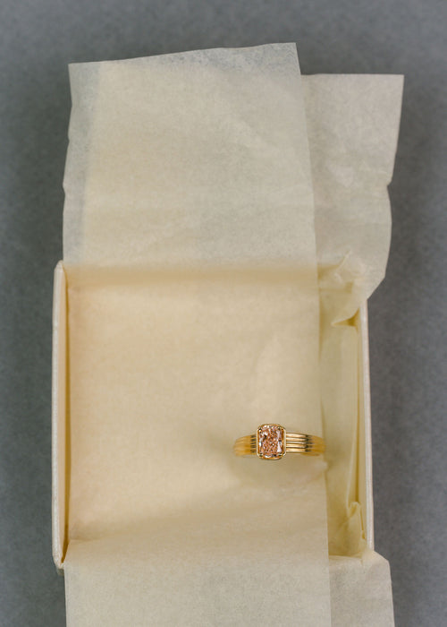 A gold ring with a rectangular pink diamond, displayed on a piece of cream-colored tissue paper. (view 5)